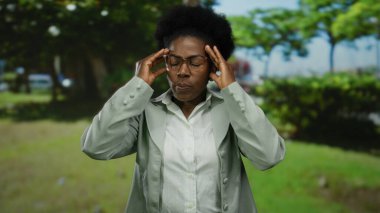 Parkta kafası tutulan bir kadın arka planda yeşillik olan bir ortamda stresli görünüyor..