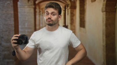 Beyaz tişörtlü adam bina koridorunda dslr kamera tutuyor, bileğinden sarkan lens kapağı; şüphecilik.