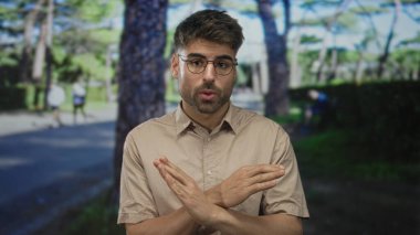 Uzun çam ağaçlarının altında yeşil parkta kollarını bağlayan bej gömlekli bir adam. Hayal kırıklığını inkar ediyor..