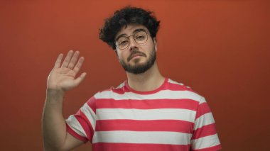 Sakallı, gözlüklü, çizgili tişörtlü, turuncu arka planda sallanan genç adam dostça selamlaşma yapıyor..