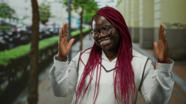 Pembe örgülü bir kadın, şehir caddesinde jest yapıyor, açık havada günlük kıyafetlerle neşe ifade ediyor, enerjik ve arkadaş canlısı ifadesiyle kişiliğini sergiliyor..