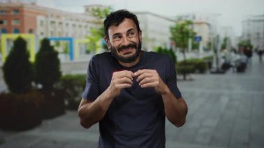 Arka planda bulanık binalar olan bir sokakta belirsizlik gösteren sakallı İspanyol bir adam..