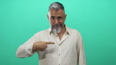 Orta yaşlı bir adam göğsünü işaret ediyor ve yeşil stüdyoda bej keten gömlekli, neşeli bir sırıtışla baş parmak işareti veriyor..