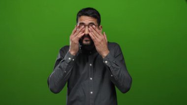 Gözlüklü, gözlerini ovuşturan genç adam yorgun olduğunu ifade eden canlı yeşil arka planda duruyor..