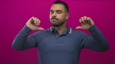 Mavi uzun kollu polo tişört giyen İspanyol genç adam iki başparmağını da morumsu stüdyoda tutuyor..