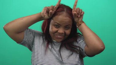 Kadın her iki işaret parmağını boynuzlara doğru kaldırır ve stüdyoda yeşil arka planda kameraya geniş bir şekilde gülümser..