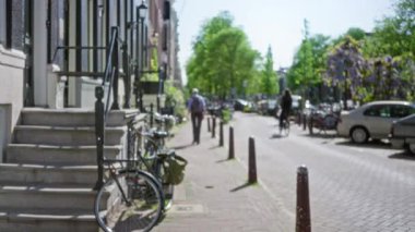 Bulanık şehir sokak sahnesinde bir adam yürüyor ve bisiklet sürüyor Avrupa 'da bir patika boyunca yeşillikler ve park edilmiş arabalar arasında güneşli bir şehir ortamında.