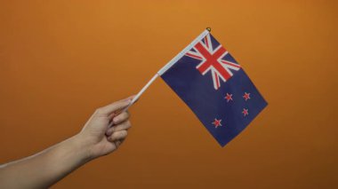 Turuncu arka planda yeni Zelanda bayrağı taşıyan adam, ulusal gurur ve kültürel kimliğini canlı renk kontrastı ve izole edilmiş erkek eli ile gösteriyor..