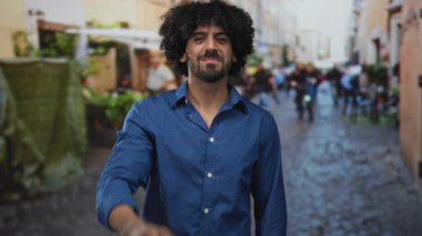 Kıvırcık saçlı, açık yakalı, göğüs ve mavi gömlekli, sokakta gülümseyen adam, insanların arkasında bulanıklaştığı samimi şehir manzarası;.