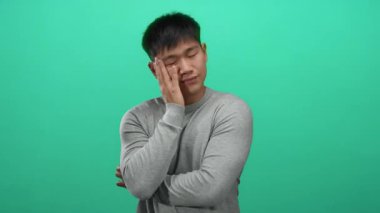 Gri kazaklı genç adam, yeşil arka plan üzerinde el ele tutuşmuş, dalgınlık ve sakinlik içinde, arkaplanda yavaşça kaynaşıyor..