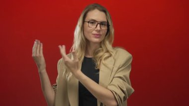 Gözlüklü ve bej ceketli, cesur kırmızı bir arkaplan karşısında elleri açık bir kadın özgüvenli ve profesyonel pozuyla dikkatleri üzerine çekiyor..