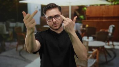 Sakallı genç adam şehir kafeteryasının terasında kaba hareketler yapıyor. Etrafı masa ve sandalyelerle çevrili restoran atmosferiyle çevrili..