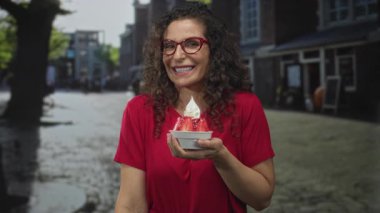 Kırmızı tişörtlü kadın bir kase çilekli tatlı tutuyor ve şehirdeki kaldırım taşı caddesinde sessizlik için parmaklarını dudaklarına işaret ediyor; sessizlik..