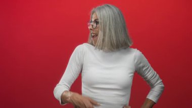 Orta yaşlı gri saçlı kadın kırmızı stüdyoda parmak göğüse gösteren beyaz uzun kollu bluz ve gözlüklü, geniş gülümsemeli, kendinden emin bir sevinç.