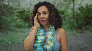 Havai zambağı olan İspanyol bikinili kadın yemyeşil bir parkta duruyor. Etrafı yeşilliklerle çevrili çeşitli duyguları gösteriyor..