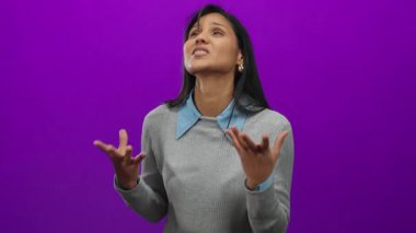 Gri bir süveter ve mavi yakalı gömleği olan canlı mor bir arka plana karşı hareketli bir kadın katı bir arkaplanda duygularını ve tarzını gösteriyor..