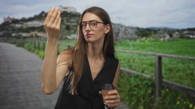 Gözlüklü ve siyah yelek giyen kadın kahve fincanını tutuyor ve kırsal kesimde tahta çit ve yeşil çitin yanında el kaldırıyor; güven daveti.