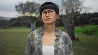 Gözlüklü genç Çinli kadın, ağaçlar ve çimlerle çevrili yeşil bir park ortamında hayal kırıklığını ifade eden bulutlu bir gökyüzünün altında derin derin düşünüyor..