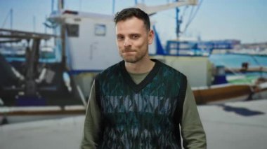 Arka planda tekneleri olan genç bir adam yeşil bir süveter giymiş, açık havada güneşli bir günde canlı mavi gökyüzünü gösterirken yakalanan yeşil bir jest yapıyor..