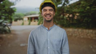 Sert bir şapka takmış genç İspanyol bir adam açık havada ağaçlarla gülüyor ve arka planda bir yol var. Açık havada sakin ama profesyonel bir anı yakalıyor..