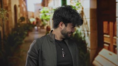 Sakallı genç adam canlı bir sokak manzarasında başını tutarak stresli ve düşünceli bir şekilde etrafı bitkilerle ve kır evleriyle çevrili duruyor..