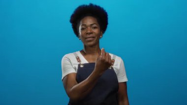 Önlüklü Afro-Amerikalı kadın kendine güvenen bir gülümsemeyle izole edilmiş mavi arka plan daveti veriyor..