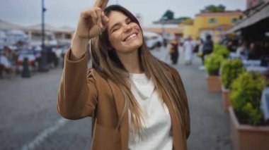 Kalabalık bir şehir caddesinde gülümseyen ve el kol hareketi yapan bir kadın. Bulanık yayalar ve canlı şehir hayatı arasında günlük kahverengi bir ceketle sıcacık bir hava yayıyor..