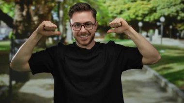 Parkta genç bir adam, siyah tişört giyiyor, başparmaklarıyla gülüyor ve kendini işaret ediyor, arka planda ağaçlar ve patikalar var, oyuncu, kendine güvenen bir tavır sergiliyor..