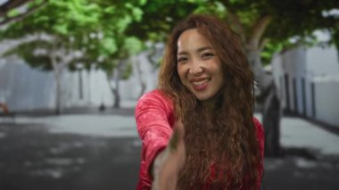 Kırmızı bluzlu kadın, yapraklı ağaçlar ve arkasında beton bina ile sokakta tokalaşmak için elini uzatıyor; dostça karşılama.