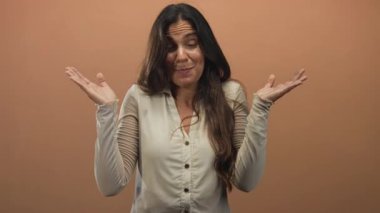 Uzun saçlı, bej bluzlu, omuz silkmiş, şeftali rengi stüdyoda kameraya bakan İspanyol bir kadın..