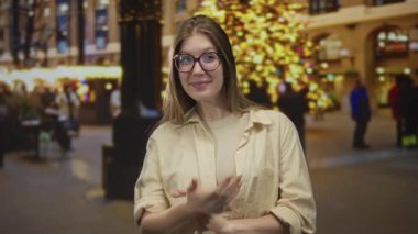 Gözlüklü kadın gülümsüyor ve parmakla işaret ediyor şehir sokaklarında gece festival ışıklarının ortasında; davet sıcaklığı.