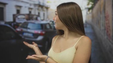 Genç kız gülüyor ve el kol hareketleri açık park halindeki arabaların arasında güneşli şehir caddesinde; neşeli karşılama daveti.