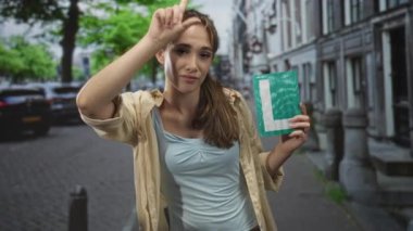 Yeşil öğrenci plakasını tutan ve kaldırımlı bir caddede park halindeki arabalarla ve kanal binalarıyla dolu bir şekilde dururken eliyle işaret eden kadın..