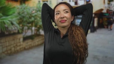 Kadın taş bir sokakta kollarını başının arkasına uzatır, yüzü açılır ve uzun kıvırcık saçlar siyah bir bluzla omuzlarının üzerine sarınır; rahatlama.