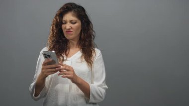 Genç İspanyol kadının gri stüdyoda iki eliyle akıllı telefonu tutarken yüzünü buruşturması..