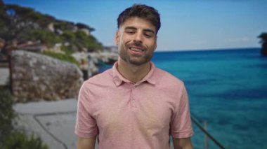 Sessiz kalmak için parmakları dudaklarına kadar uzanan adam güneşli bir sahilde hafif dalgalarla pembe bir polo gömlek giyiyor..