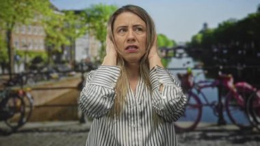 Güneşli bir günde kalabalık bir şehirde dar bir kanal sokağının yanında kederli bir ifadeyle kulakları kaplayan kadın; gürültü sıkıntısı.