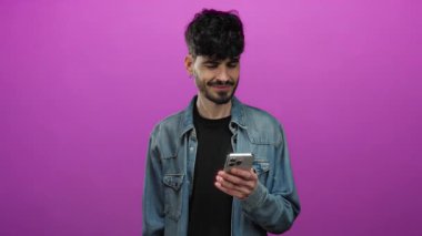 Akıllı telefonuyla gülümseyen genç adam izole edilmiş pembe arka planda kot ceket giyiyor.