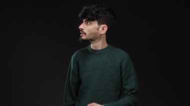 Yeşil kazaklı genç adam, şaşkın bir ifadeyle karanlık arka plana işaret ediyor merak ve entrika taşıyor..