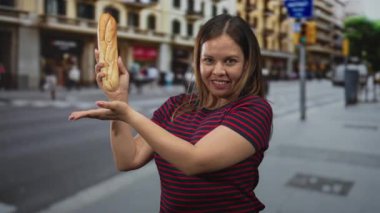 Elinde baget tutan ve sokağın ortasında başparmağını kaldıran bir kadın gülümseyerek ekmeği sunuyor; dostça bir onay.