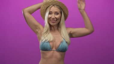 Mavi bikinili bir kadın, pembe stüdyoda çıplak göbeğini gösteren kollarını kaldırmış hasır bir şapka takıyor..