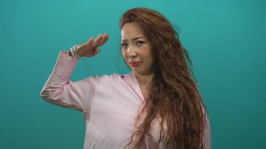 Genç bir kadın, pembe bluz ve uzun dalgalı saçlar giyerek bir deniz mavisi stüdyosunda el pençe divan selam veriyor..