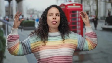 Kırmızı telefon kulübesinin önünde el kol hareketi yapan ve arka planda bulanık yayalar ve ağaçlar olan şehir caddesinde renkli bir kazak giyen bir kadın..