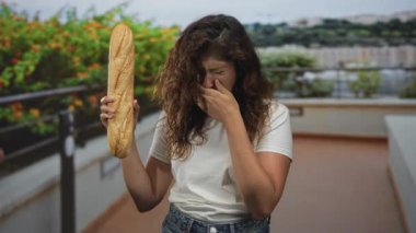 Elinde bir baget tutan ve burnunu çimdikleyen bir bina korkuluğu olan bir kadın saksı bitkileri ve uzak şehir manzaralı bir bina;.