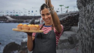 Bir kadın elinde ahşap bir kek tepsisiyle sahil taşı basamaklarında kamerayı işaret ediyor; konukseverliği sıcak karşılıyor.