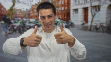 Şehirli bir sokakta başparmağıyla gülümseyen genç adam canlı bir şehirde mutluluk ve pozitiflik sergiliyor..