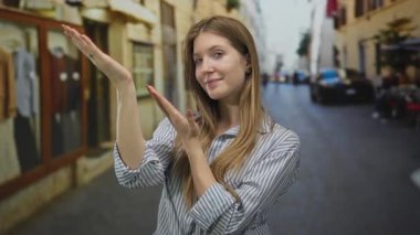 Sarışın kadın, yaz ışığı altında, çizgili gömlek ve uzun saç çerçeveli mağaza önü sahnesi giyerek avuçlarını kaldırıyor..