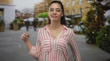 Gözlüklü, çizgili tişörtlü genç İspanyol kadın hareketli caddede başparmak kaldırıyor; iyimserliği destekliyor.