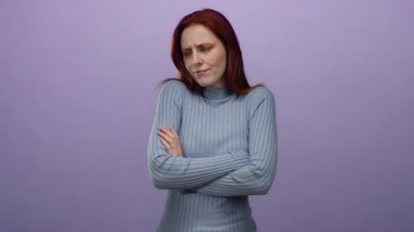 Kızıl saçlı kadın, kollarını mor arka plana doğru uzatmış düşünceli bir duruşta, hafif ışıkta durgun bir ifade sergiliyor, mavi kazak giyiyor ve sükuneti taşıyor.