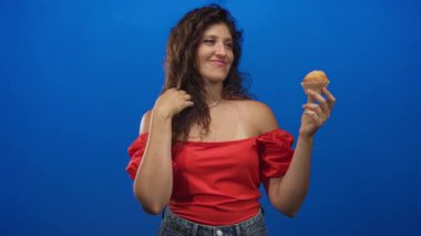 Kadın bir elinde kek tutuyor ve stüdyoda çıplak omzunu gösteren saçlara dokunuyor..